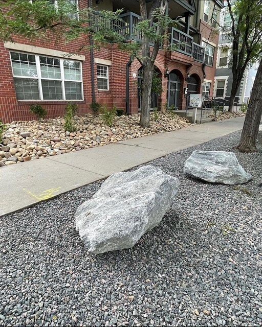 Two large gray rocks in a gravel landscape beside a sidewalk and brick building