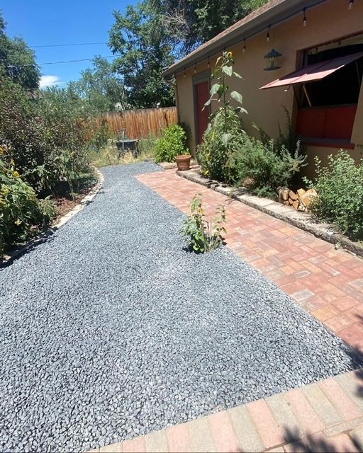 Gravel path beside a brick house with potted plants, shrubs, and a sunny garden walkway