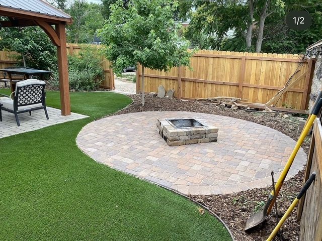 Backyard patio with a circular paver fire pit, green lawn, wooden fence, and covered seating area