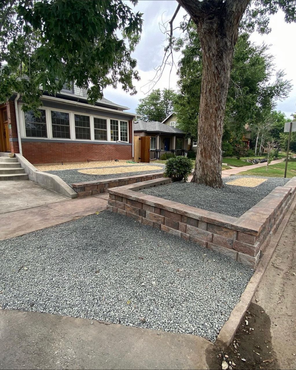 Landscaped front yard with gravel beds, large tree, and a brick house with steps and windows
