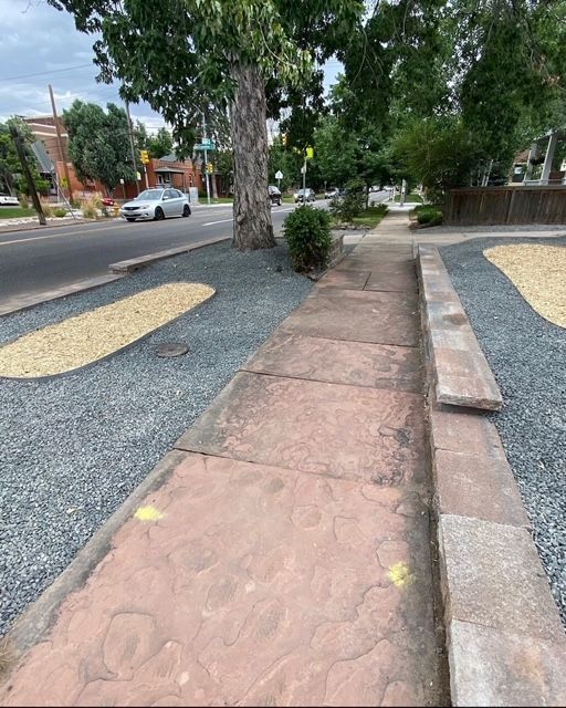 Cracked sidewalk with tree roots, bordered by gravel, beside a quiet street and parked cars.